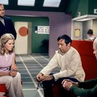 Der Text "RECEPTION" ist in einem Bereich mit Schalter zu sehen. Vier Menschen sitzen und stehen in einem Raum mit futuristischer Einrichtung.