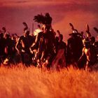 Ich kenne den Text nicht. Eine Gruppe von Menschen mit Federschmuck bewegt sich durch ein goldenes Feld bei Sonnenuntergang.