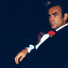 Eine Person mit ernstem Blick in Anzug mit roter Blume im Knopfloch, dunkler Hintergrund, stark hervorgehoben.