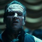 Ein Mann mit verschlossenem Gesichtsausdruck, Narben auf der Stirn, vor einem unscharfen, gewellten Hintergrund.