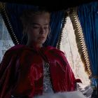 Eine Frau in einem prächtigen roten Umhang sitzt in einem Raum mit blauen, reich verzierten Vorhängen.