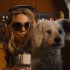 Eine Frau mit Kopfhörern und großer Sonnenbrille sitzt neben einem weißen Hund in einem Cockpit.