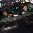 Zwei Männer sitzen in einem Auto, einer hält eine Waffe. Sie schauen angespannt in unterschiedliche Richtungen.