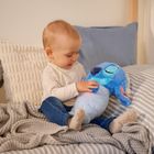Ein Baby sitzt auf einem Bett mit gestreiftem Bezug, hält ein blaues Plüschtier und ist von Kissen und Decken umgeben.