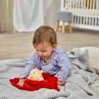 Kein Text vorhanden. Ein Kleinkind spielt auf einer grauen Decke mit einem Plüschtier. Im Hintergrund ist ein Kinderbett.