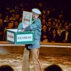Text: "ICE CREAM". Ein Mann in blauer Jacke und heller Mütze trägt drei Eiscremekisten in einer Arena mit Publikum.