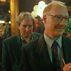Zwei Männer in einem festlichen Raum, der vordere lächelt mit einem Getränk in der Hand. Beide tragen Anzüge, umgeben von Partygästen.