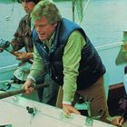 Drei Menschen auf einem Boot, zwei Männer und eine Frau in Outdoor-Kleidung. Einer angelt, die anderen zwei schauen konzentriert.