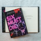 Buchcover: "SIX MUST DIE", Victoria Wlosok. Neon-Schrift mit traurigem Smiley, zersplittert. Bücher im Hintergrund.