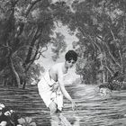 Keine erkennbaren Texte. Illustration: Eine Frau kniet im flachen Wasser eines Waldbachs, umgeben von Blumen und Bäumen.