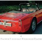 Nummernschild: DA-HM 466H. "Triumph TR4" über dem Autoheck. Rotes Cabrio auf gepflasterter Straße, Natur im Hintergrund.