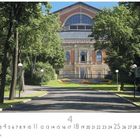 Bayreuth, Festspielhaus. Ein Weg führt durch Bäume zu einem großen Gebäude mit Blumenbeeten und Verkehrsschildern.