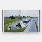 Ein offenes Buch zeigt eine architektonische Szene mit einem modernen, dreieckigen Gebäude in grüner Umgebung.