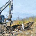 Ein Bagger mit der Aufschrift "Volvo" hebt Erde. Eine Person in Schutzkleidung steht daneben auf einem unbebauten Feld.