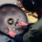 Person bearbeitet ein Hangs. Sie hält Werkzeuge an der Trommel. Tisch mit Werkzeugen im Hintergrund.