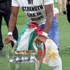 Person mit weißem T-Shirt "MY STRENGTH LIES IN JESUS" hält große silberne Trophäe auf einem Sportplatz.