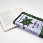 Auf einem Tisch liegt ein aufgeschlagenes Buch. Daneben liegt "Frankenstein" von Mary Shelley als Reclam-Ausgabe.