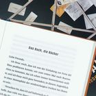 Ein aufgeschlagenes Buch zeigt den Titel "Das Buch, die Bücher" und beginnt mit "Liebe Freunde,". Im Hintergrund sind Briefe.