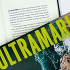 „ULTRAMARIN“ in großen gelben Buchstaben über einem Foto von Wellen, das über einer Buchseite mit schwarzem Text liegt.