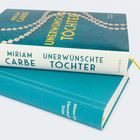 Miriam Carbe, Unerwünschte Töchter. Zwei blau-grüne Bücher, eines geöffnet mit Perlenmuster, das andere geschlossen.