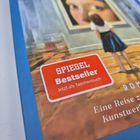 "SPIEGEL Bestseller", "Jetzt als Taschenbuch", "ROMAN". Kind betrachtet großes Gemälde in einem Raum.