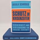 Buchcover mit Text: „Ursula Schröder, Schutz in Krisenzeiten, Sicherheit und Verteidigung für unsere Zukunft“, Farben: blau und orange.