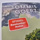 "TOMMIE GOERZ", roter Sticker mit "SPIEGEL Bestseller-Autor", Hintergrund: Landschaft mit Bäumen und Wolken.