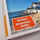 Text: "SPIEGEL Bestseller-Autor" auf rotem Hintergrund. Szene: Sandstrand mit Häusern und Meer im Hintergrund.