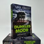 Der Text zeigt "Lars Engels, Dunkles Moor, Janosch Janssen ermittelt, Ullstein". Ein hölzernes Haus steht in einer düsteren Landschaft.