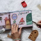 Ein geöffnetes Kinderbuch zeigt eine Illustration: Ein Gnomenkapuzenmantel mit der Zahl "2" sitzt, umgeben von winterlichem Dekor.