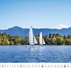 Kalendariumsmonat April. Zwei Segelboote auf einem See, dahinter bewaldete Hügel und Berge unter einem klaren Himmel.