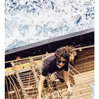 04: Eine Person klettert eine Netztreppe auf einem Schiff hoch, umgeben von Holzplanken und schäumendem Meer.