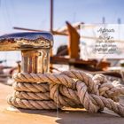 Text: „Aufbruch. Ein Schiff im Hafen ist sicher, doch dafür werden Schiffe nicht gebaut.“ – John Augustus Shedd. Dargestellt ist ein Metallpoller mit Seil, im Hintergrund Boote im Hafen.
