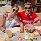 Zwei Personen an einem Holztisch mit rustikaler Jause, Getränken und Sonnenschein, in gemütlicher Atmosphäre.