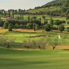 Grüne Hügellandschaft mit Golfplatz, Bäumen und einem Gebäude im Hintergrund. Eine weiße Fahne markiert das Loch.
