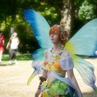 Person in buntem Elfenkostüm mit großen, blauen Flügeln und Blumenkranz, spaziert im sonnigen Park.