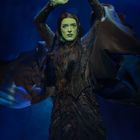 Eine Frau mit grünem Gesicht und erhobenen Armen, in einem dunklen, fließenden Kleid vor blauem Hintergrund.