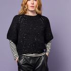 Eine Person mit rotblondem Haar trägt ein schwarzes Oberteil, darunter ein gestreiftes Shirt, kurze Lederhose, vor lila Hintergrund.