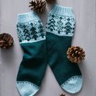 Zwei gestrickte Socken in Dunkelgrün und Hellblau mit Tannenbaummuster, umgeben von Tannenzapfen auf grauem Hintergrund.