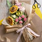 Eine Blumensträuße aus gehäkelten Blumen, darunter Sonnenblumen und Rosen, in braunem Papier mit rosa Schleife gebunden. Im Hintergrund eine Schere.