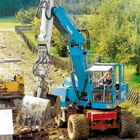Blauer Bagger mit der Aufschrift "FUCHS". Der Bagger hebt Erde auf einer Baustelle in ländlicher Umgebung.