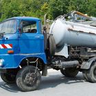 Ein blauer Lastwagen mit einem silbernen Tankaufbau steht im Freien. Der Hintergrund zeigt Bäume und Gebäude.