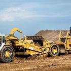 Die gelben Baumaschinen mit "CATERPILLAR 088 631E" bewegen Erde auf einer Baustelle vor einem Erdhügel und blauem Himmel.
