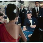 Personen in Abendkleidung sitzen an einem Spieltisch. Im Fokus ist ein Mann im Smoking in einem Casino.