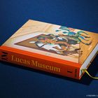 Auf einem blauen Hintergrund liegt ein Buch mit dem Titel "Lucas Museum" auf dem Rücken.
