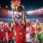 "April" und Kalender angezeigt. Ein Spieler in rotem Trikot hält jubelnd einen Pokal, umgeben von Teamkollegen. Stadion im Hintergrund.