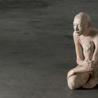 Text: "Tijenodli". Eine sitzende Skulptur einer nachdenklichen, nackten Figur auf grauem Boden.