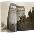 Text "HANOMAG" auf einem Industriegebäude. Vier Männer sitzen auf einer Mauer, unterhalten sich. Umliegende Straßenszene.