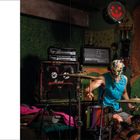 Drummer mit bunter Maske und Kopfhörern spielt Schlagzeug in einem Raum mit Verstärkern und bunten Tapeten. Links Textblock.
