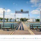 Text: "Heiligendamm". Eine lange Holzbrücke führt über blaues Wasser zu weißen Gebäuden vor einem bewaldeten Hintergrund.